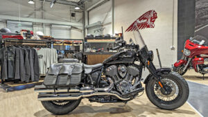 Une moto Indian noire avec sacoches est exposée à l'intérieur d'une salle d'exposition, avec du matériel et des accessoires d'équitation sur des étagères et des rayonnages à l'arrière-plan.