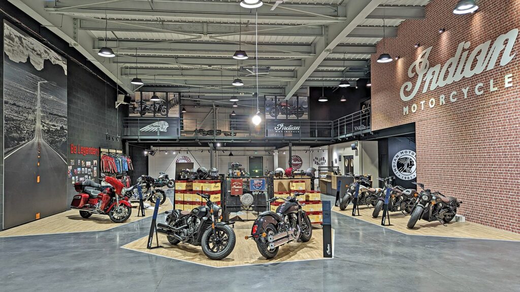 Une salle d'exposition de motos moderne présente diverses Indian Motorcycles, des accessoires et des produits de marque dans un décor de style industriel et avec de grands logos muraux.