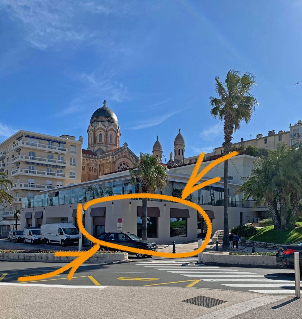 Un bâtiment en bord de mer avec de grandes fenêtres et des tours d'église en forme de dôme à l'arrière-plan ; un cercle orange et une flèche indiquent le rez-de-chaussée et la zone d'entrée.