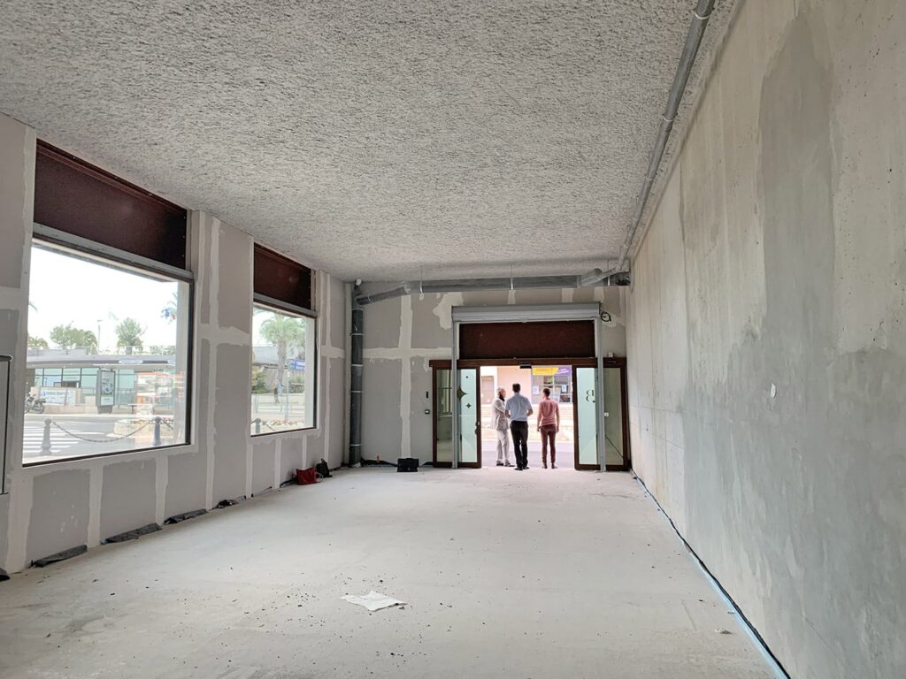 Un espace commercial vide et inachevé, aux murs nus et aux grandes fenêtres ; trois personnes se tiennent près des portes d'entrée ouvertes, tournées vers l'extérieur.