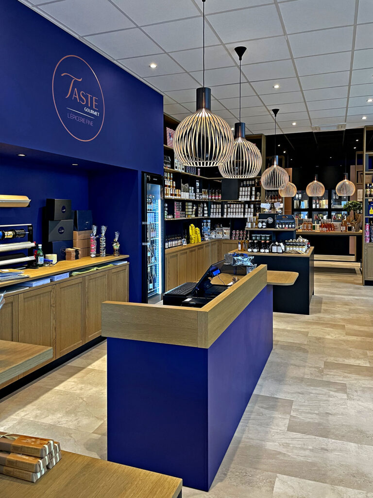 Intérieur d'une épicerie fine moderne avec des étagères en bois, des accents bleus, des lampes suspendues et des présentoirs de produits alimentaires et de bocaux.