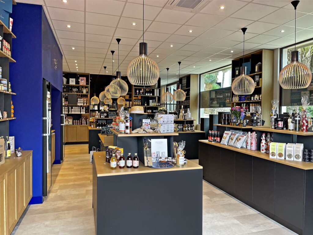 Intérieur d'un magasin moderne de produits alimentaires spécialisés, avec des étagères et des présentoirs contenant divers produits gastronomiques, des bouteilles et des articles de cadeaux, sous des lampes suspendues décoratives.