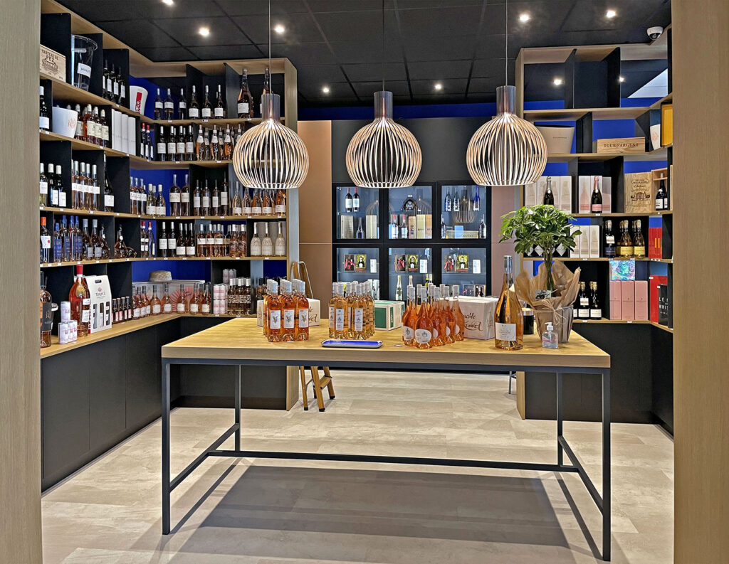 Intérieur d'un magasin de vin moderne avec des étagères de bouteilles assorties, une table présentant du vin et des cadeaux, et trois lampes suspendues décoratives suspendues au-dessus.