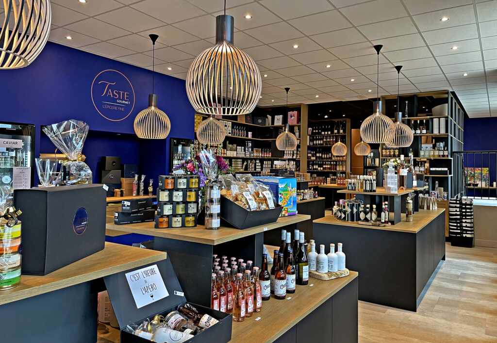 Intérieur d'une épicerie fine moderne avec des étagères et des comptoirs en bois présentant divers produits alimentaires, des boissons et des paniers-cadeaux sous des lumières suspendues.