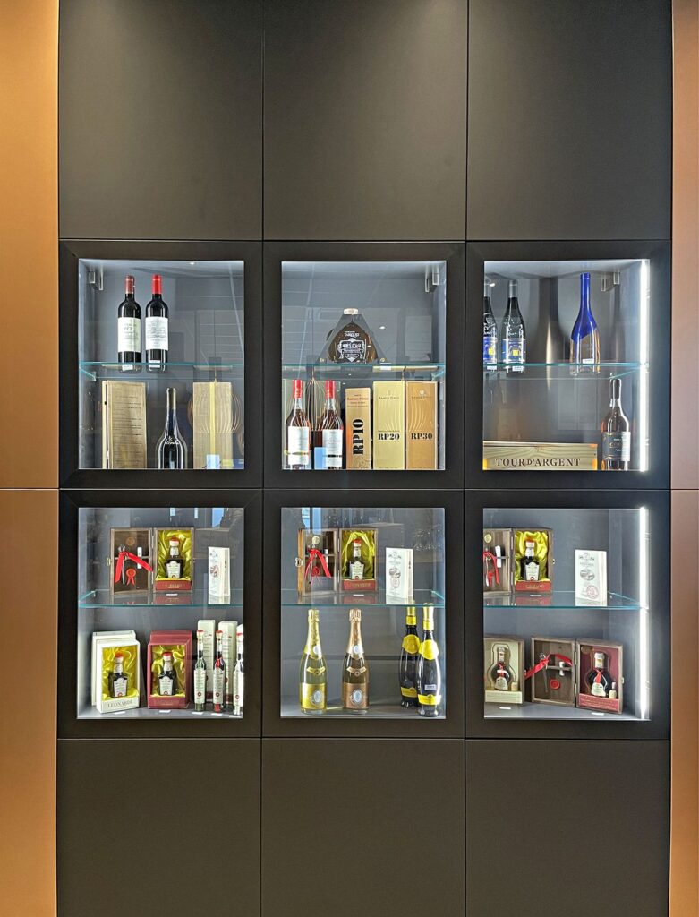 Un présentoir mural avec des étagères en verre contenant diverses bouteilles de vin et de liqueur, des boîtes en bois et des objets encadrés, disposés dans six compartiments éclairés.