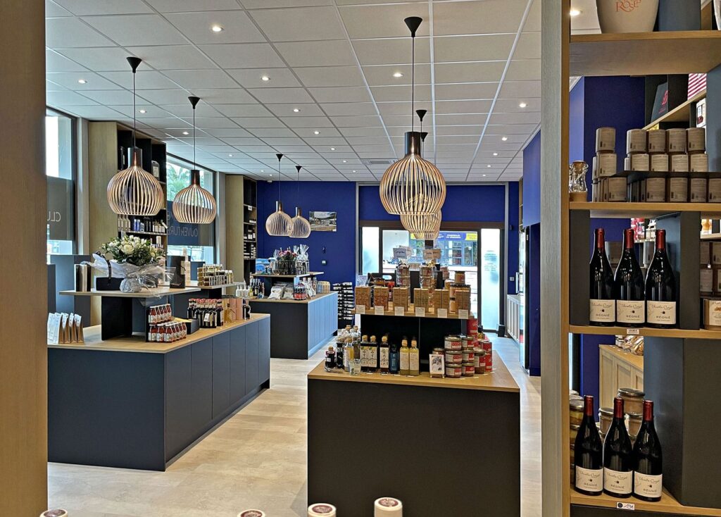 Intérieur d'une épicerie fine moderne avec des étagères et des comptoirs présentant divers produits alimentaires, des bocaux, des bouteilles et du vin, le tout sous des lampes suspendues et entouré de murs d'accentuation bleus.