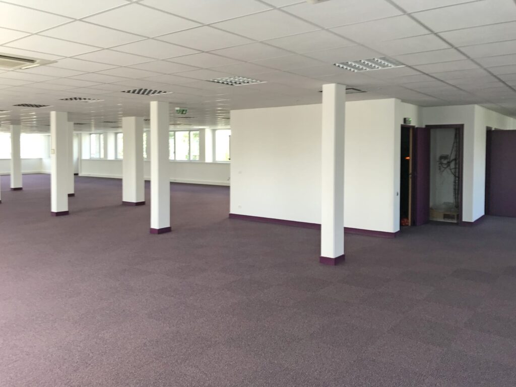 Grand espace de bureau vide aux murs blancs, à la moquette violette, aux multiples piliers et à l'éclairage fluorescent. Plusieurs portes et fenêtres sont visibles le long du mur du fond.