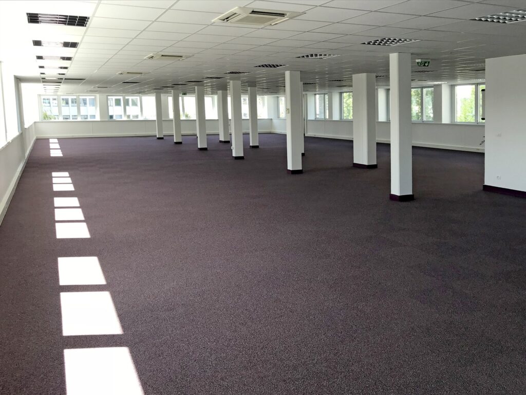 Grand espace de bureau vide avec un sol recouvert de moquette, des murs blancs, des plafonniers et de multiples colonnes de soutien ; des fenêtres s'alignent sur le côté gauche pour laisser entrer la lumière naturelle.