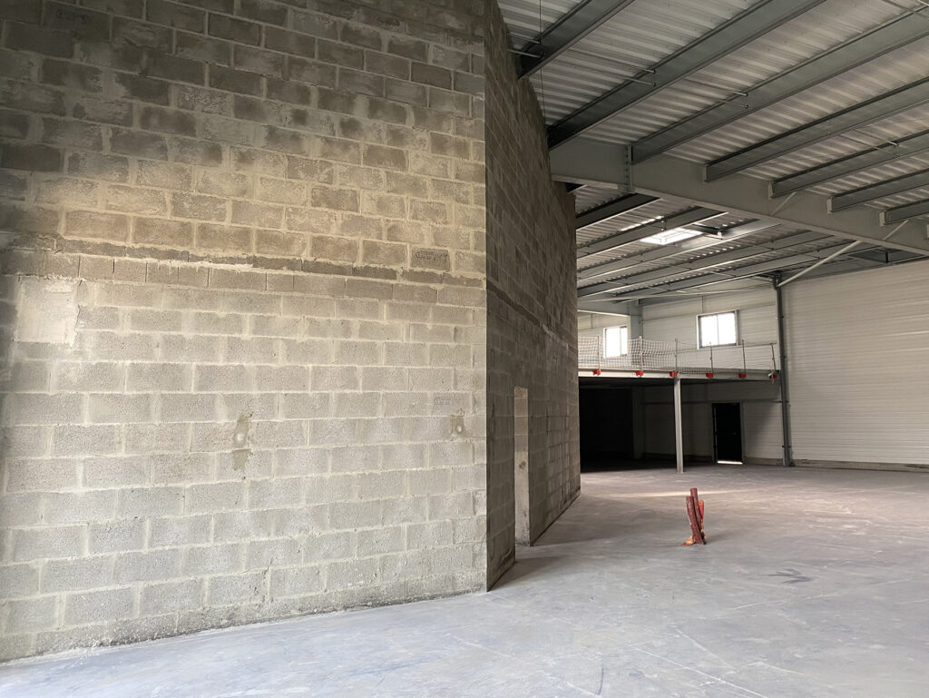 Intérieur d'un entrepôt industriel inachevé avec des murs en blocs de béton, des poutres métalliques apparentes et des tuyaux visibles au sol.