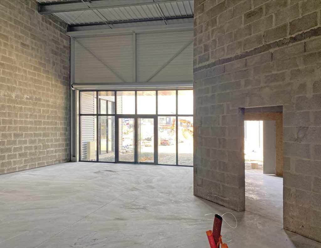 Espace industriel vide avec des murs en béton, de grandes fenêtres et une petite porte ; des matériaux de construction sont visibles à l'extérieur.