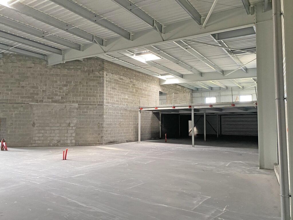 Intérieur d'un entrepôt vide avec des murs en béton, un haut plafond métallique et quelques bornes de sécurité rouges. La lumière naturelle pénètre par plusieurs fenêtres.