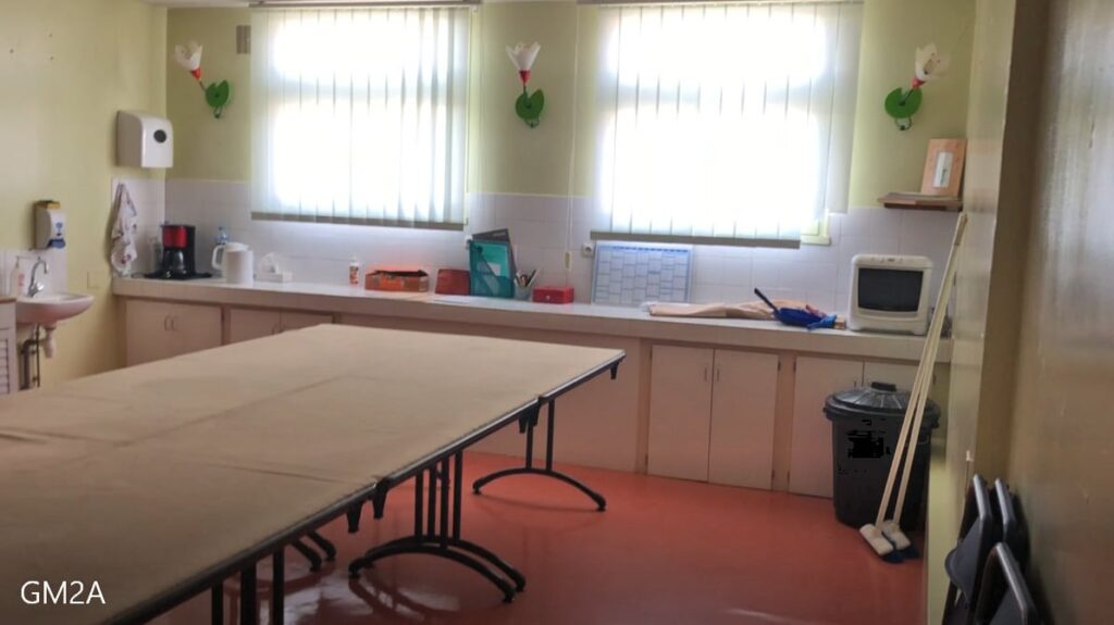 Une kitchenette avec une longue table, des armoires, un évier, de petits appareils électroménagers, des produits de nettoyage et deux fenêtres avec des stores qui laissent entrer la lumière naturelle.
