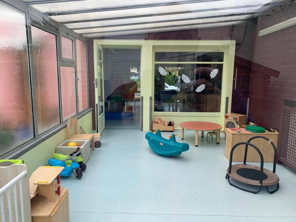 Salle de jeux intérieure avec divers jouets, dont une balançoire à bascule, des véhicules autoportés, une table ronde et un mini-trampoline ; de grandes fenêtres laissent entrer la lumière naturelle.