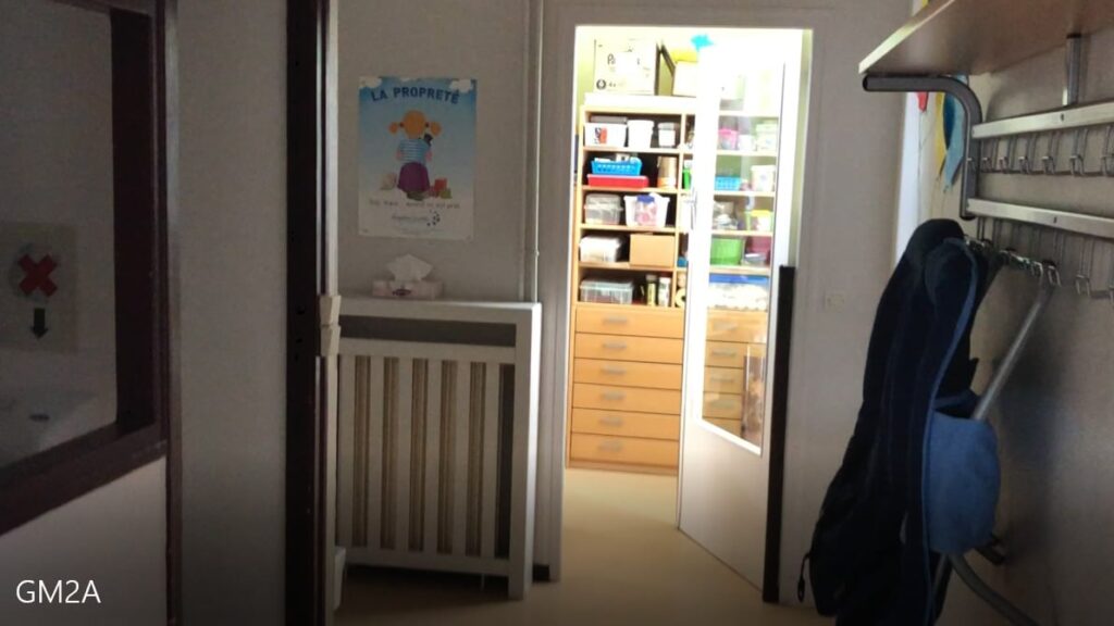 Un couloir avec des patères, un berceau, un poster sur le mur et une porte ouverte révélant des étagères avec des fournitures organisées dans une pièce bien éclairée.