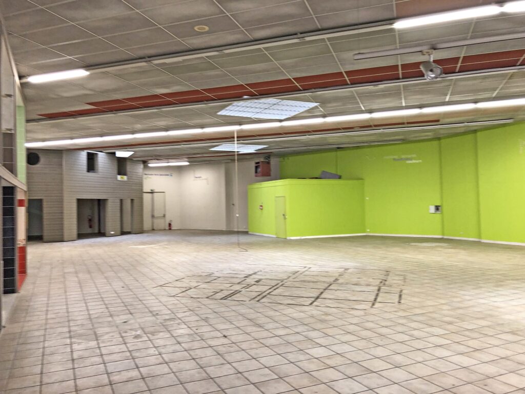 Grande pièce vide avec un sol carrelé, des murs verts et beiges et des panneaux de plafond apparents. Une corde pend du plafond et des portes de maintenance sont visibles.