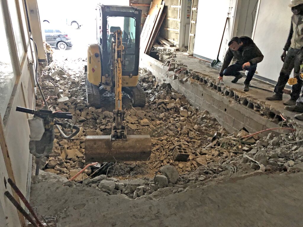 Une petite pelleteuse travaille à l'intérieur d'un bâtiment, brisant le sol en béton, tandis que deux personnes observent le chantier.