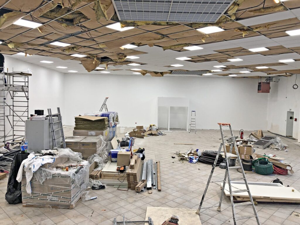 Chantier de construction avec des matériaux épars, des échafaudages et des échelles. Le plafond est partiellement exposé, montrant l'isolation et les luminaires.