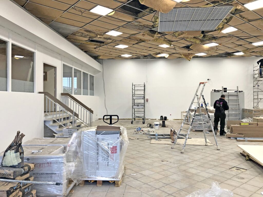 Une grande pièce en cours de rénovation avec des panneaux de plafond apparents, des matériaux de construction, des échelles et un ouvrier sur place.