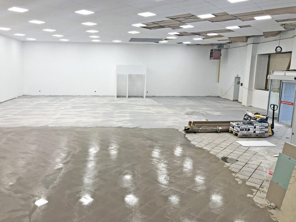 Grande pièce intérieure en cours de rénovation avec un sol inachevé, des piles de matériaux de construction et des dalles de plafond partiellement enlevées.