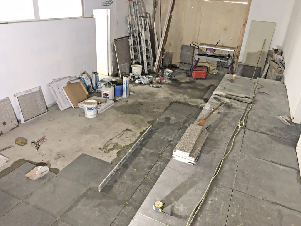 Une pièce en cours de rénovation avec des outils, des carreaux et des matériaux de construction éparpillés sur le sol ; des carreaux sont partiellement installés le long d'un côté.