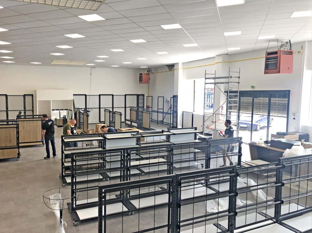Plusieurs personnes assemblent et organisent des étagères métalliques dans un grand espace commercial bien éclairé, avec de hauts plafonds et des matériaux de construction éparpillés.