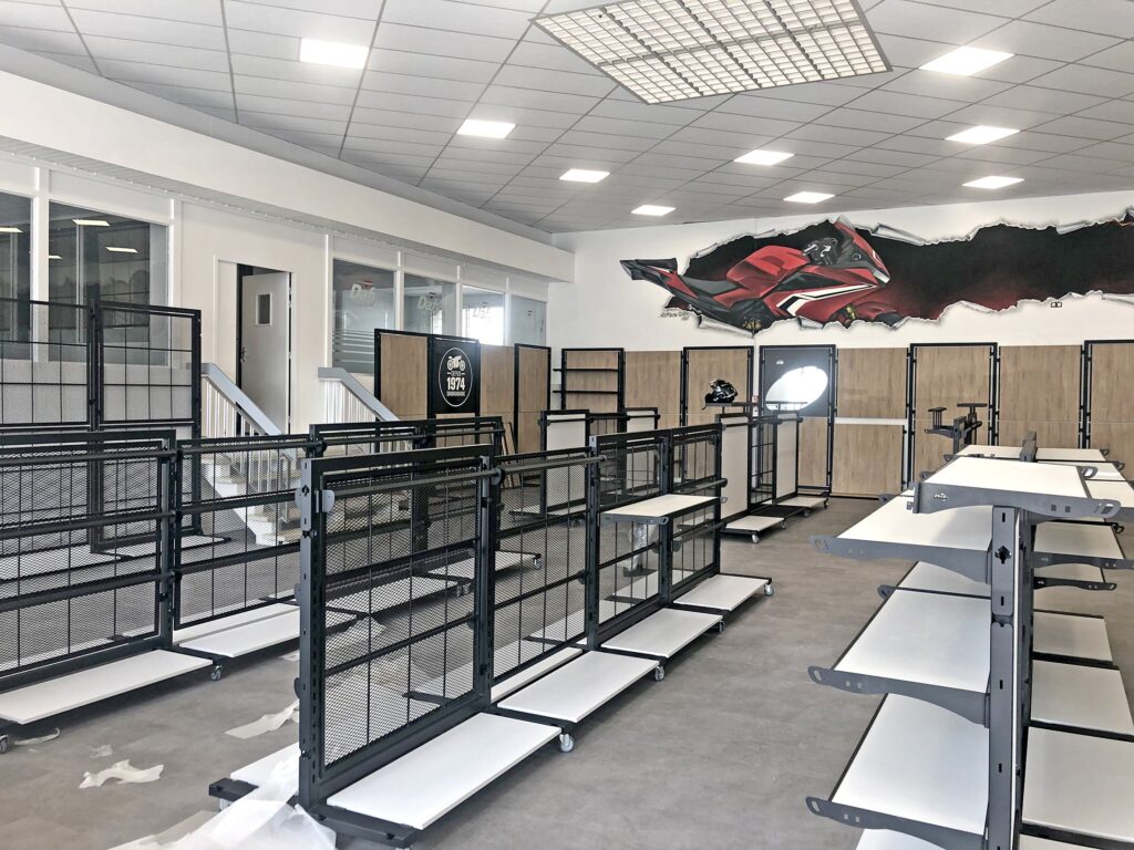 Des étagères et des rayonnages vides en métal noir sont disposés dans un espace commercial très éclairé, avec une peinture murale et des panneaux de bois à l'arrière-plan.