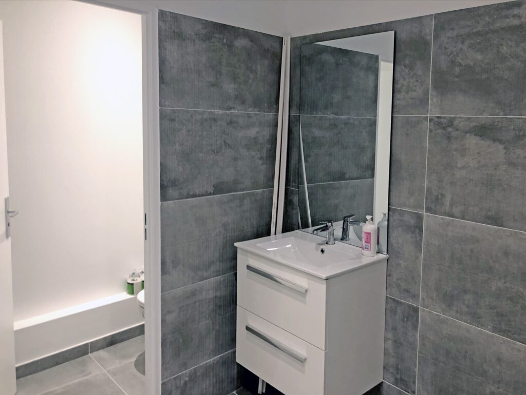 Salle de bains moderne avec des murs carrelés gris, un meuble-lavabo blanc avec un évier, un miroir et des articles de toilette ; une porte ouverte révèle une vue partielle des toilettes et de la baignoire.