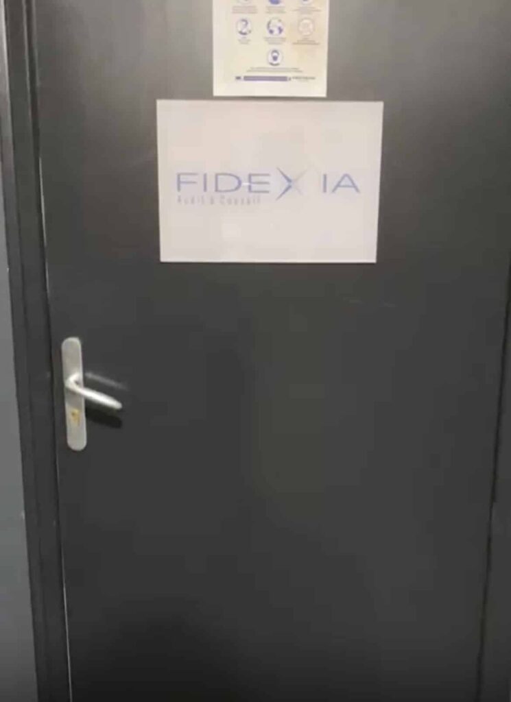 Une porte grise fermée avec une poignée argentée et un panneau portant l'inscription "FIDEXIA" en lettres bleues. Un petit panneau avec des icônes est affiché au-dessus du panneau principal.