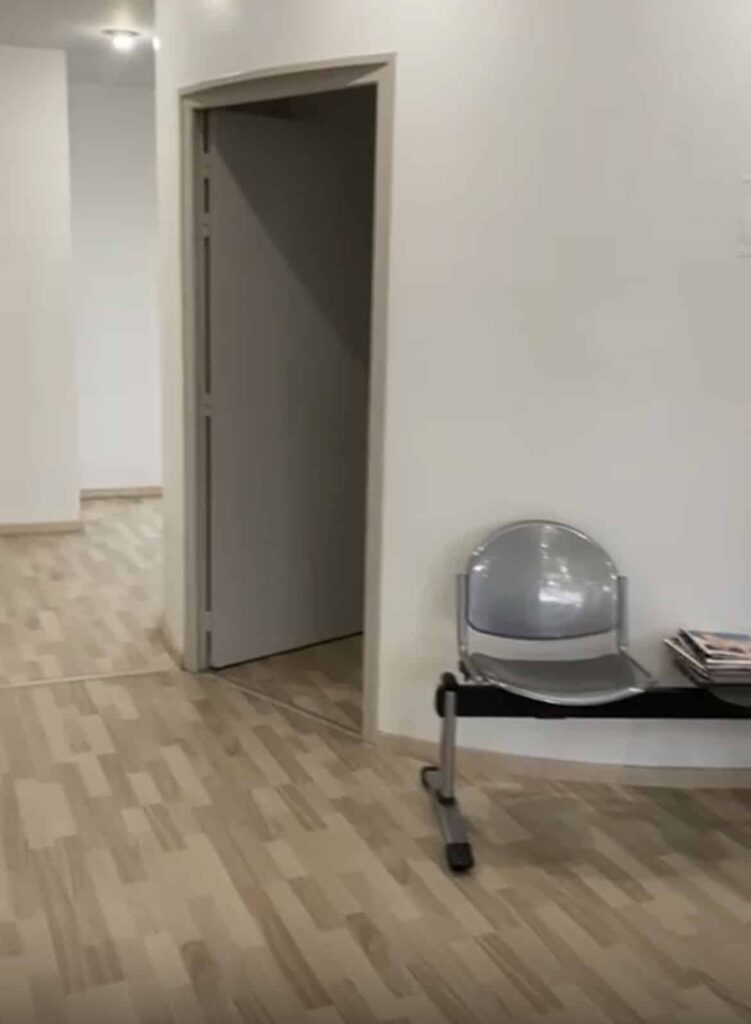 Une salle d'attente avec une chaise métallique, une pile de magazines, un sol en bois clair et une porte ouverte menant à une autre pièce.