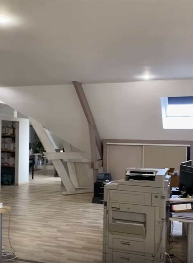 Espace de bureau avec plafond incliné, imprimante, bureau, ordinateur, étagères et plancher en bois ; la lumière du jour passe par une petite fenêtre.
