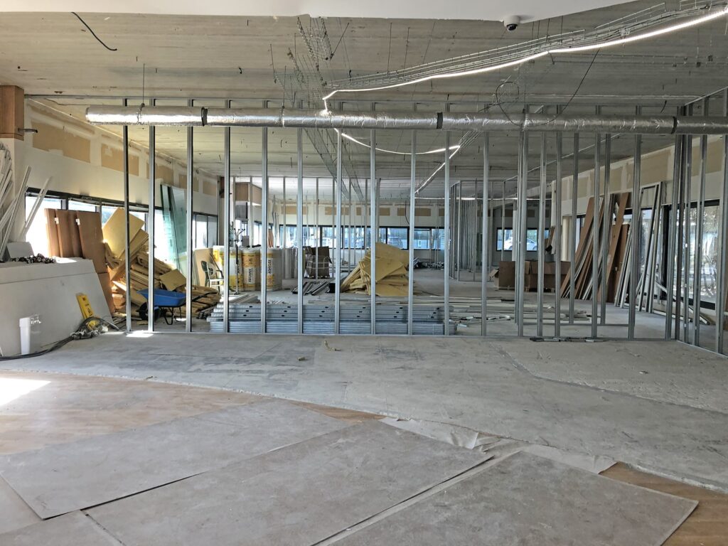 Grand espace intérieur en cours de construction avec des murs à ossature métallique apparente, des matériaux de construction éparpillés, des outils et des sols non finis.