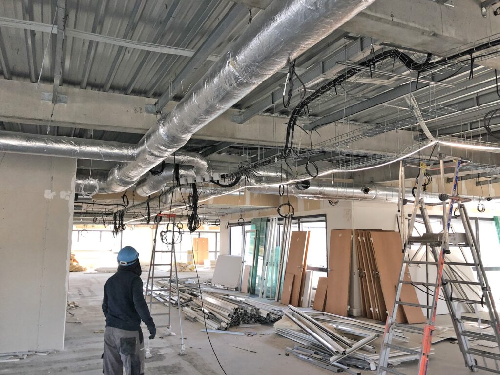 Un ouvrier de la construction se tient dans un bâtiment inachevé où sont exposés des conduits, des câbles électriques, des échelles et des matériaux de construction éparpillés sur le sol.