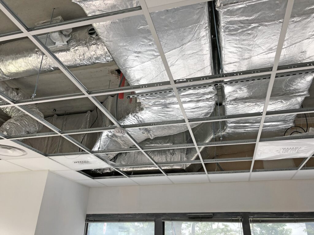Un plafond suspendu auquel il manque plusieurs dalles laisse apparaître les gaines, les tuyaux et l'isolation d'un local commercial ou d'un bureau.