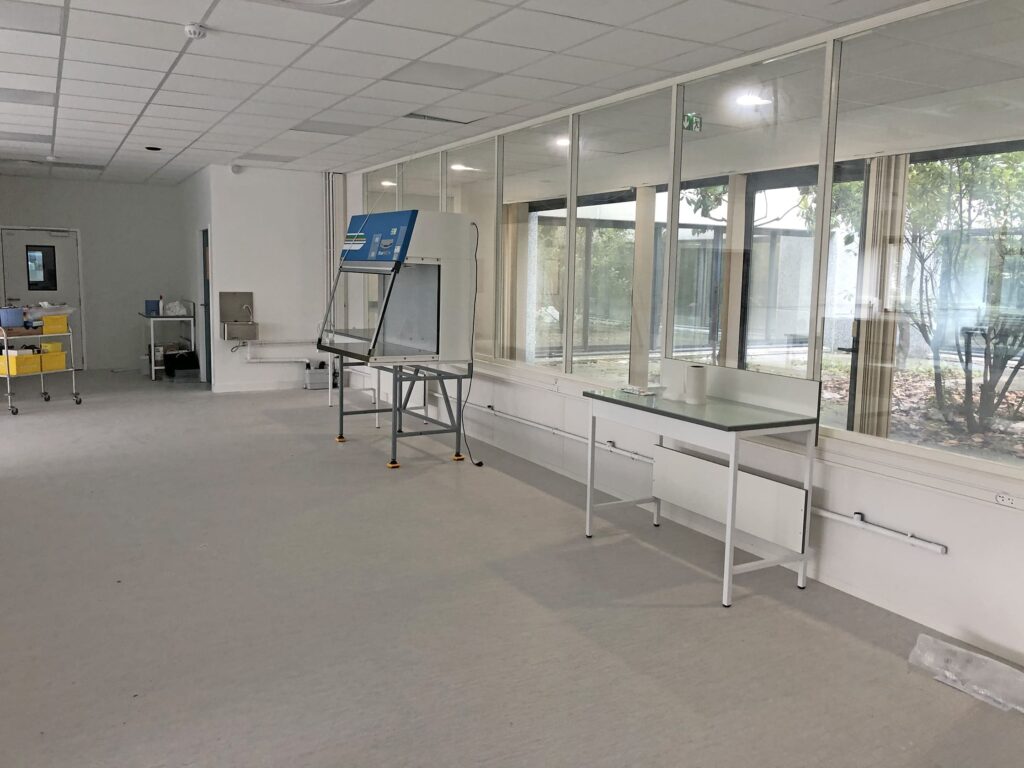Une salle de laboratoire pratiquement vide avec une armoire de sécurité biologique, des tables et de grandes fenêtres donnant sur les arbres d'une cour extérieure.