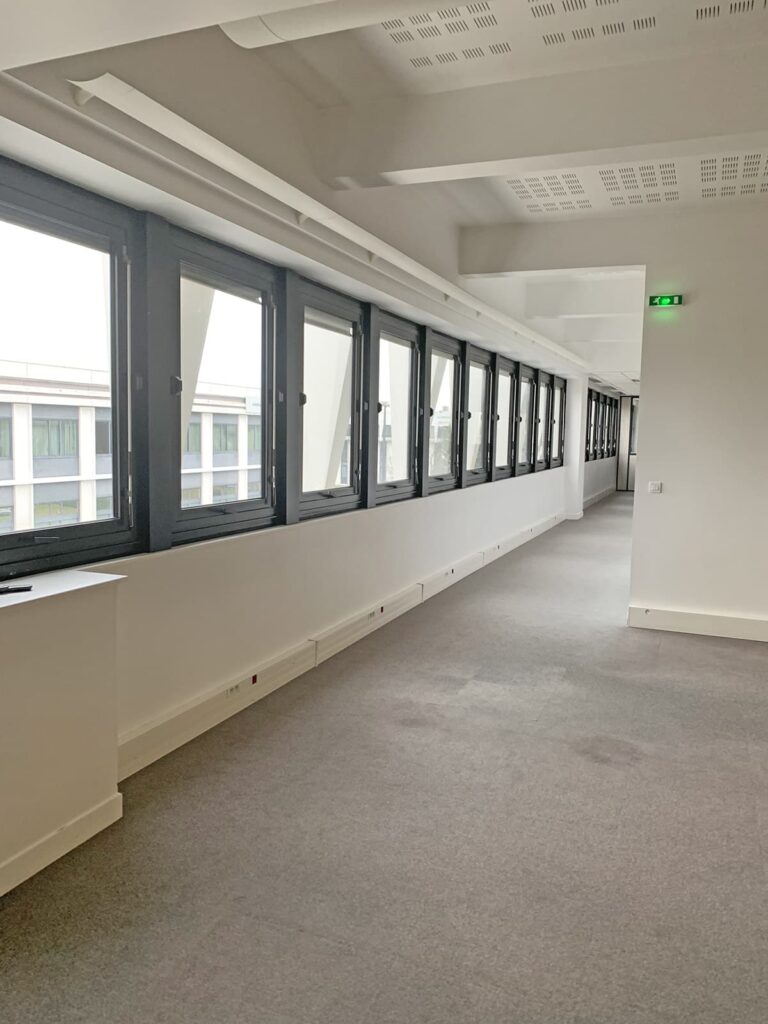 Un long couloir vide avec une moquette grise, de grandes fenêtres d'un côté, des murs blancs et un panneau de sortie vert à l'extrémité.