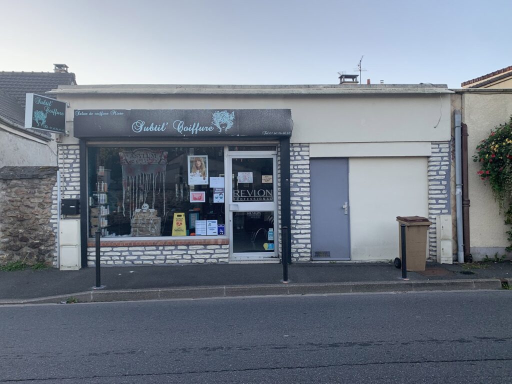 Petit salon de coiffure avec une enseigne noire indiquant "Subtil' Coiffure", une vitrine en verre, des affiches dans la fenêtre et une porte latérale grise. Le bâtiment est de couleur claire avec des accents de pierre.
