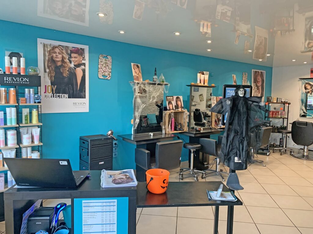 Un salon de coiffure avec des murs bleus, des présentoirs de produits capillaires, des décorations d'Halloween, notamment une sorcière suspendue et un seau à citrouille, et des postes de coiffure avec des chaises et des miroirs.