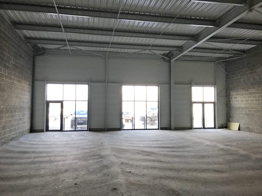 Entrepôt industriel vide avec sol en béton, haut plafond métallique et trois grandes fenêtres du sol au plafond laissant entrer la lumière du jour ; plusieurs voitures garées sont visibles à l'extérieur.