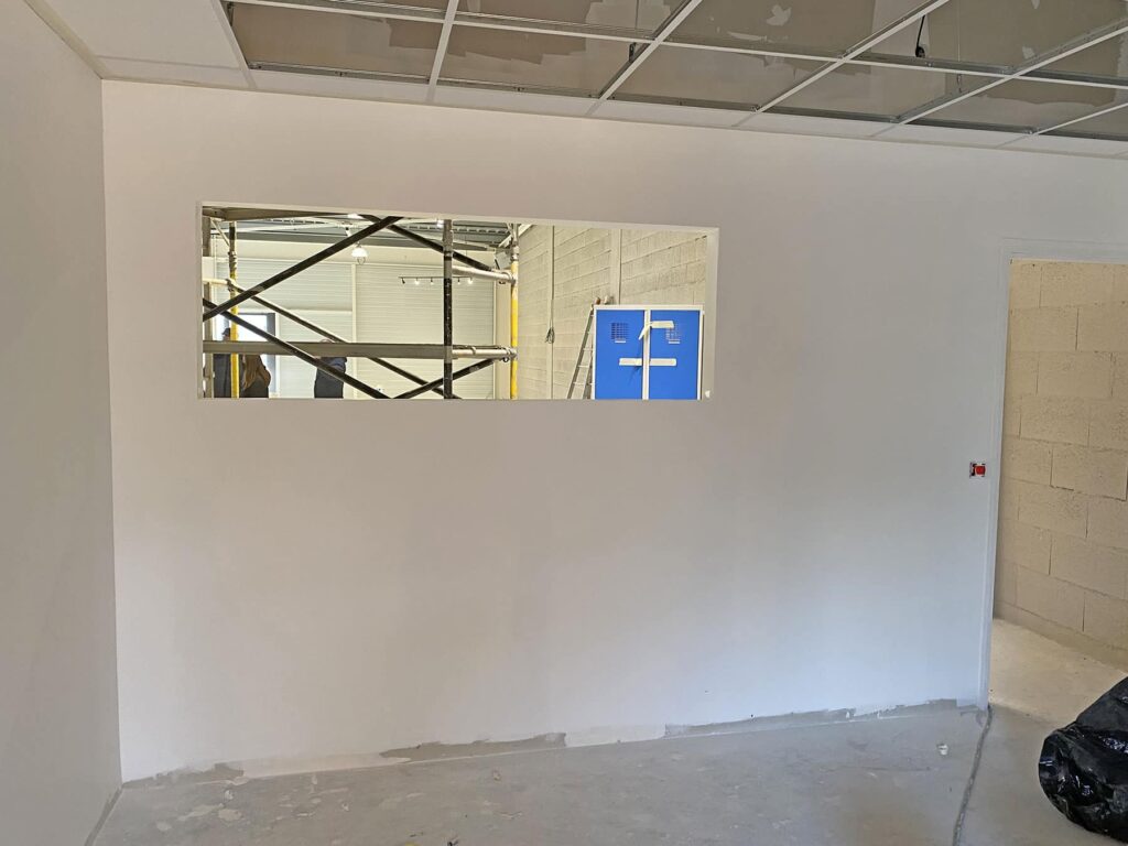 Mur intérieur blanc avec une ouverture de fenêtre rectangulaire, montrant des échafaudages et des équipements bleus dans une pièce inachevée au-delà ; les dalles de plafond sont partiellement installées.