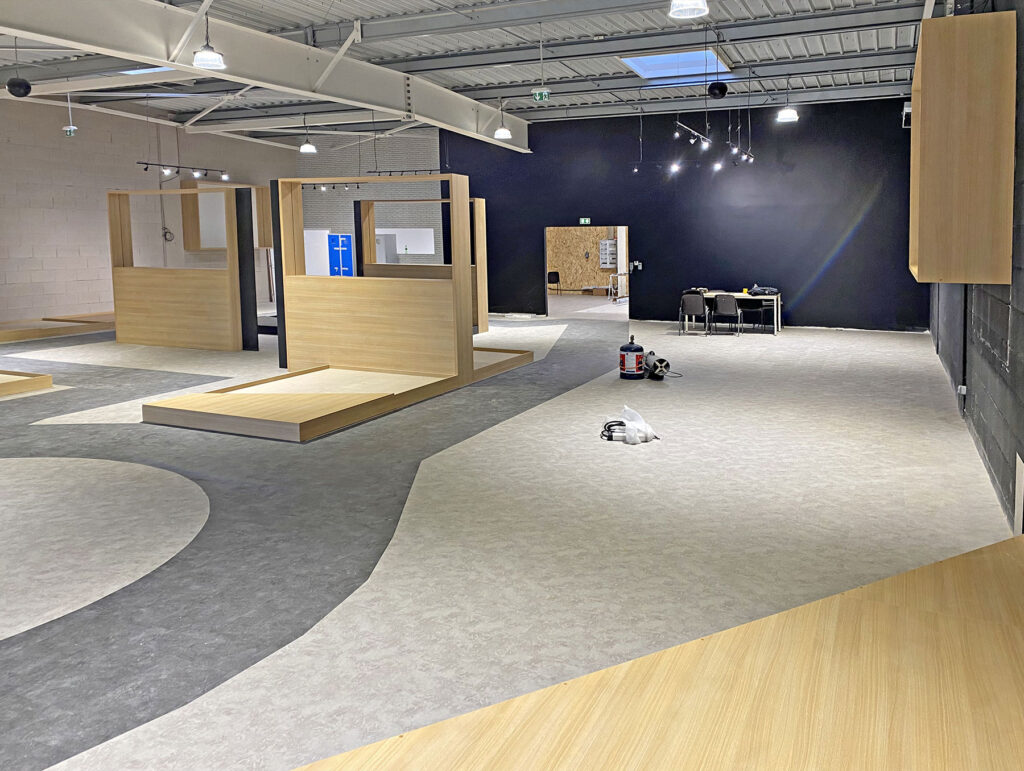 Intérieur d'un espace commercial moderne et inachevé avec des plateformes d'exposition en bois clair, des murs sombres, un plafond apparent et des matériaux de construction sur le sol.