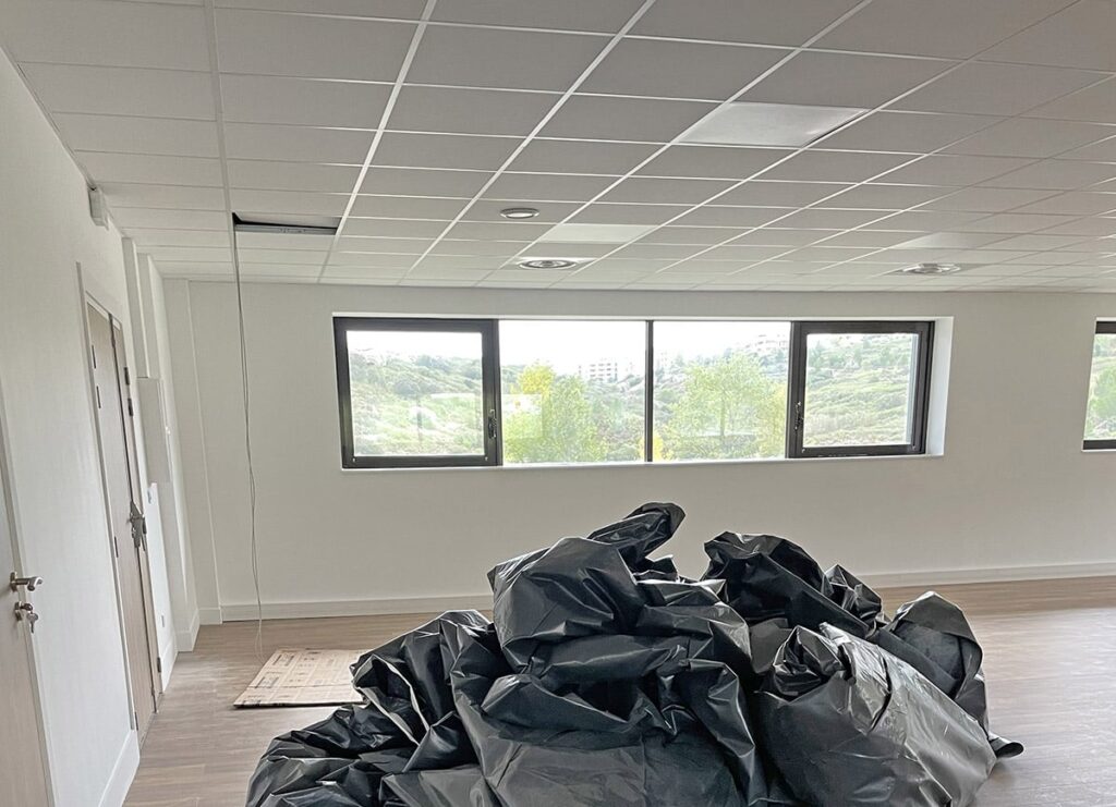 Une pile de sacs poubelles noirs est posée sur le sol d'une pièce vide aux murs blancs, aux dalles de plafond et aux trois fenêtres donnant sur un paysage verdoyant.