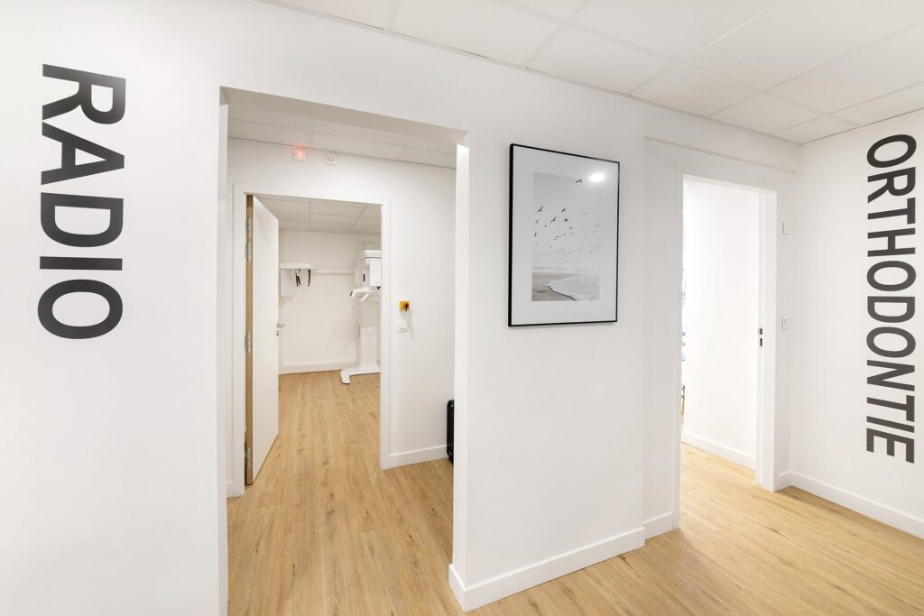 Un couloir de clinique dentaire moderne avec des sols en bois, des murs blancs et des panneaux indiquant "RADIO" et "ORTHODONTIE" à côté des portes ouvertes.