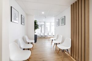 Une salle d'attente moderne et lumineuse avec des chaises blanches alignées le long des deux murs, un sol en bois clair, des œuvres d'art encadrées et une plante en pot.