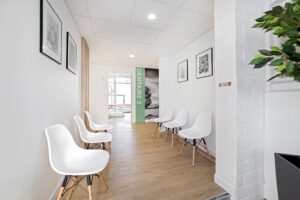 Une salle d'attente propre et moderne avec des chaises blanches le long des murs, un sol en bois et des œuvres d'art en noir et blanc. Un panneau indiquant "DENTAIRE 2" est visible au bout du couloir.