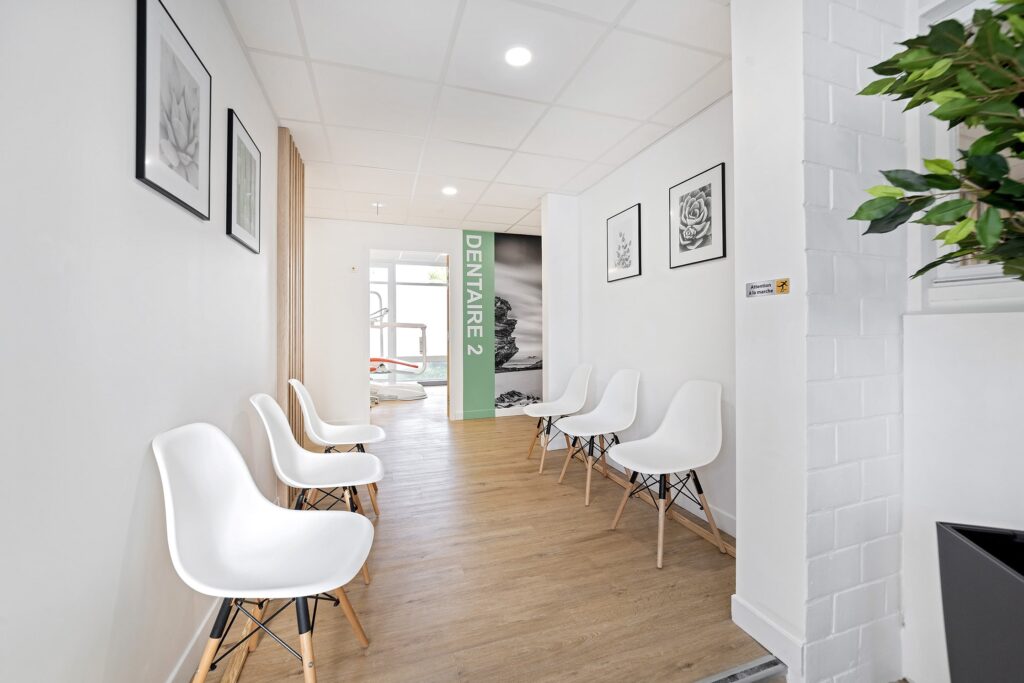 Une salle d'attente propre et moderne avec des chaises blanches le long des murs, un sol en bois, des photos encadrées et un panneau vert indiquant "DENTAIRE 2" au bout du couloir.