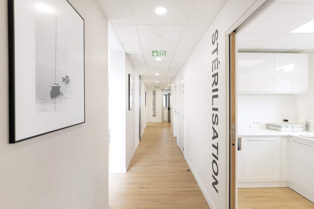 Un couloir propre dans une clinique médicale ou dentaire avec des panneaux "Stérilisation" et "Orthodontie", des murs blancs, un sol en bois et une photo encadrée de bateaux.