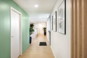 Un couloir propre avec un sol en bois clair, des murs verts et blancs, des œuvres d'art encadrées, des plantes en pot et une vue sur une salle de réception lumineuse.
