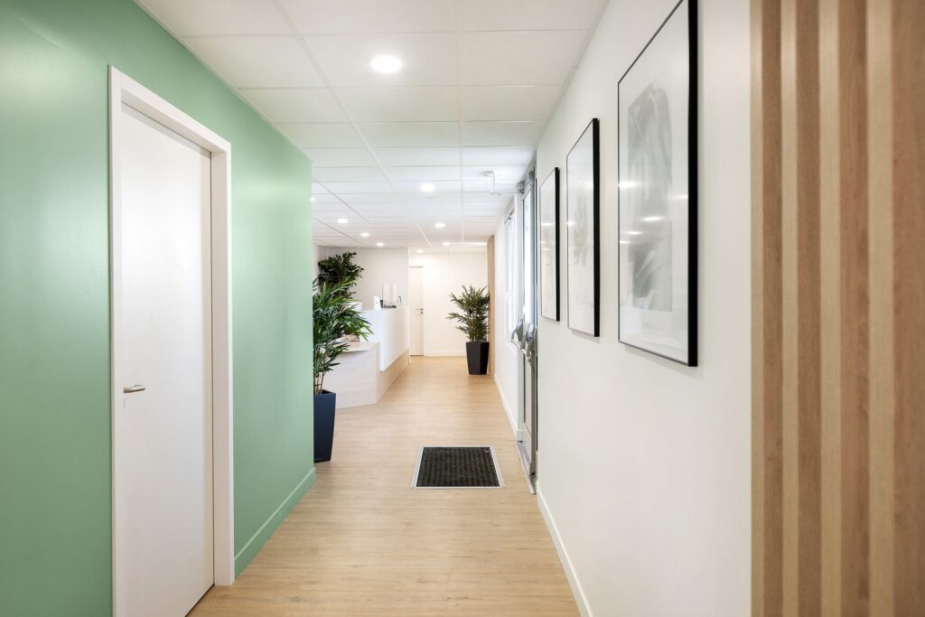 Un couloir moderne avec un sol en bois clair, des murs verts et blancs, des œuvres d'art encadrées, des plantes en pot et une porte blanche. Le couloir mène à une salle de réception lumineuse.