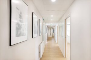 Un couloir propre et bien éclairé avec des œuvres d'art encadrées sur le mur de gauche, du parquet et des portes menant à des chambres ; un panneau indiquant "ORTHODONTIE" est visible à l'extrémité.