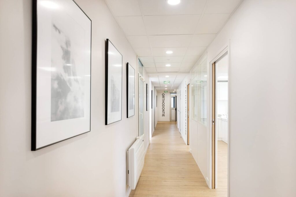 Couloir moderne et lumineux avec un sol en bois clair, des murs blancs, des œuvres d'art encadrées et des portes menant à des bureaux ; un panneau indiquant "ORTODONTIE" est visible au bout du couloir.